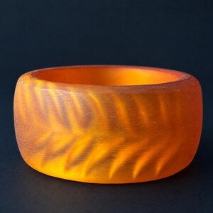 May include: A wide, translucent orange bangle bracelet featuring a repeating, etched chevron or leaf-like pattern across its exterior surface. The material has a matte, frosted finish that allows light to pass through, creating depth and colour variation.