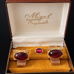 May include: A vintage gold-coloured cufflink and tie tack set with large oval, deep red cabochon stones. The set is presented in a brown box with the name "Migel Originals" printed on the inside lid.