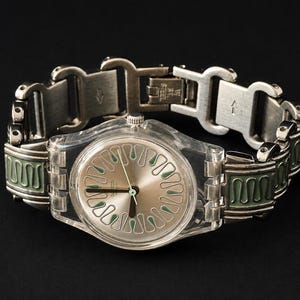 May include: A silver-toned wristwatch with a clear plastic case and a silver dial featuring green accents. The watch has a silver metal band with green enamel detailing. The watch is displayed against a black background.