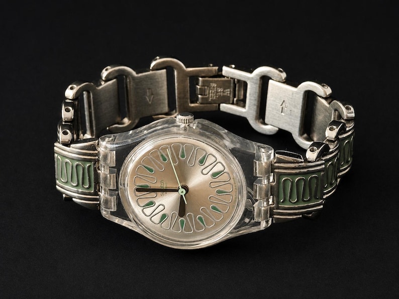 May include: A silver-toned wristwatch with a clear plastic case and a silver dial featuring green accents. The watch has a silver metal band with green enamel detailing. The watch is displayed against a black background.