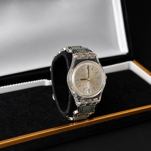 May include: A clear wristwatch with a silver-colored face and a transparent band with green accents. The watch is displayed in a black box with a gold trim and a black velvet interior.