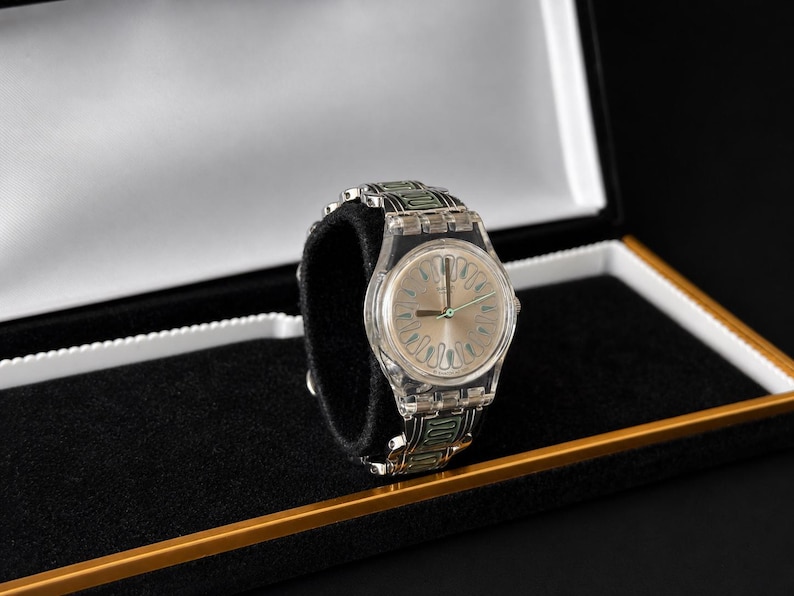 May include: A clear wristwatch with a silver-colored face and a transparent band with green accents. The watch is displayed in a black box with a gold trim and a black velvet interior.