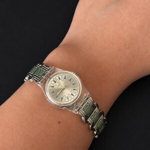 May include: A silver-toned wristwatch with a clear plastic case and a green and silver band. The watch face is silver with a unique design. The watch is worn on a wrist.