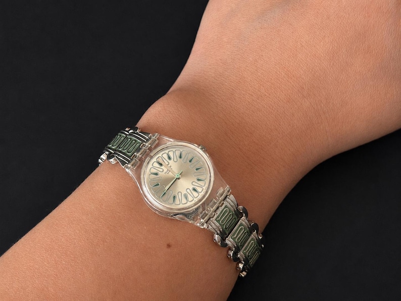May include: A silver-toned wristwatch with a clear plastic case and a green and silver band. The watch face is silver with a unique design. The watch is worn on a wrist.