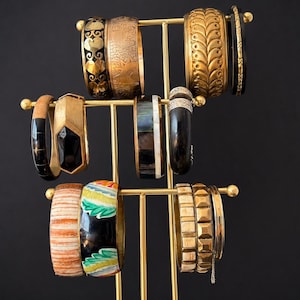 Peut inclure: Un présentoir à bijoux en métal doré portant une collection de divers bracelets rigides. Les bracelets présentent des designs variés incluant des découpes en cœur, des motifs gravés, des incrustations noir et blanc et des motifs floraux peints.