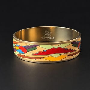 May include: A gold-toned bangle bracelet with a colorful geometric design. The bracelet features a repeating pattern of abstract shapes in red, blue, green, and yellow. The interior of the bracelet is a solid gold color, and the text "FREY WILLE" is visible.