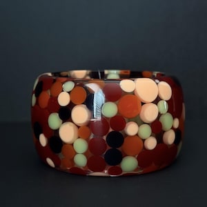 May include: A wide bangle bracelet made of clear resin embedded with numerous circular discs in varying sizes. The embedded circles feature a color palette of deep burgundy, dark brown, pale peach, and soft mint green throughout the piece.