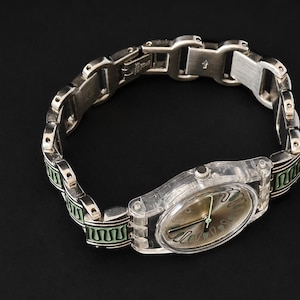 May include: A silver-toned wristwatch with a clear plastic case and a metallic band. The watch face is a muted gold color with black hands. The band features green and black decorative accents.