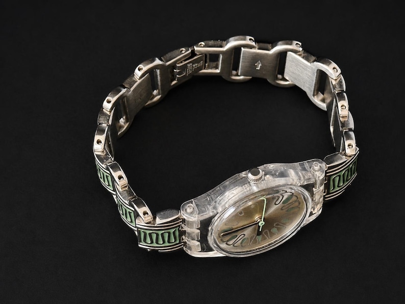 May include: A silver-toned wristwatch with a clear plastic case and a metallic band. The watch face is a muted gold color with black hands. The band features green and black decorative accents.