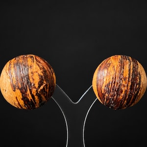 May include: A pair of round, spherical earrings featuring a textured surface with a marbled pattern of dark brown and light tan streaks. The earrings are displayed on a clear, Y-shaped stand against a solid black background.