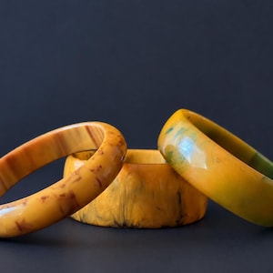 May include: Three circular bangle bracelets with smooth, polished surfaces. One bracelet features a translucent amber tone with dark brown mottled patterns, while the other two display solid yellow and green marbled resin textures.