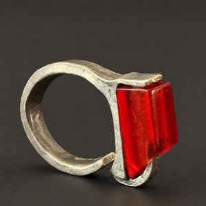 May include: A ring featuring a thick, textured, silver-toned metal band with a matte finish. The band holds a single, rectangular, translucent red stone set vertically at the front, creating a bold and industrial appearance with visible metal work.