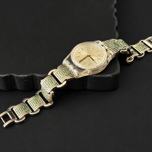 May include: A gold-toned wristwatch with a transparent plastic case and a gold dial. The watch band features a repeating pattern of gold and light green rectangular links. The watch is displayed on a dark surface.
