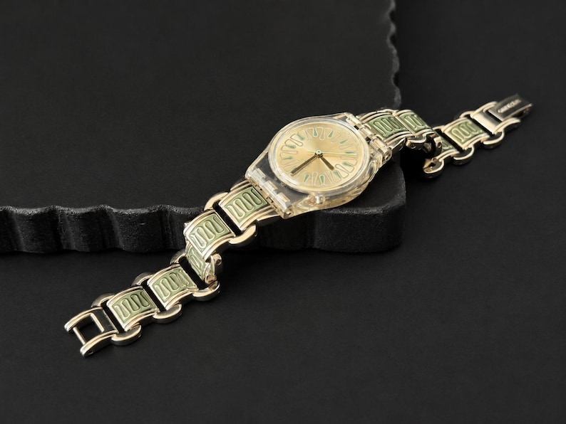 May include: A gold-toned wristwatch with a transparent plastic case and a gold dial. The watch band features a repeating pattern of gold and light green rectangular links. The watch is displayed on a dark surface.