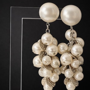 May include: A pair of elegant, off-white pearl earrings. Each earring features a large, round pearl at the top, connected by a silver chain to a cluster of smaller pearls and silver leaf-shaped accents. The background is black.