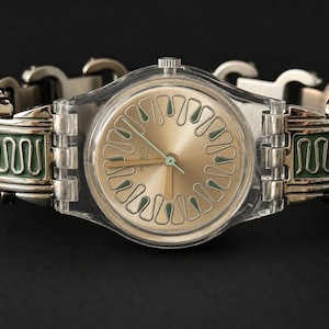 May include: A Swatch watch with a transparent plastic case and a gold-colored face. The watch has a silver-toned metal band with green and silver decorative elements. The watch hands are gold-colored.