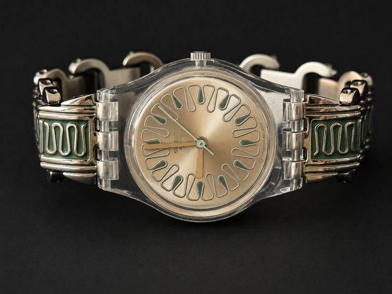 May include: A Swatch watch with a transparent plastic case and a gold-colored face. The watch has a silver-toned metal band with green and silver decorative elements. The watch hands are gold-colored.