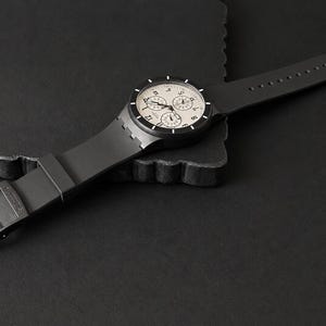 K&ouml;nnte beinhalten: Eine schwarze Armbanduhr mit hellem Zifferblatt und drei Hilfszifferbl&auml;ttern. Die Uhr hat ein schwarzes Armband und einen Schnallenverschluss. Die Uhr wird auf einer dunklen Oberfl&auml;che pr&auml;sentiert.