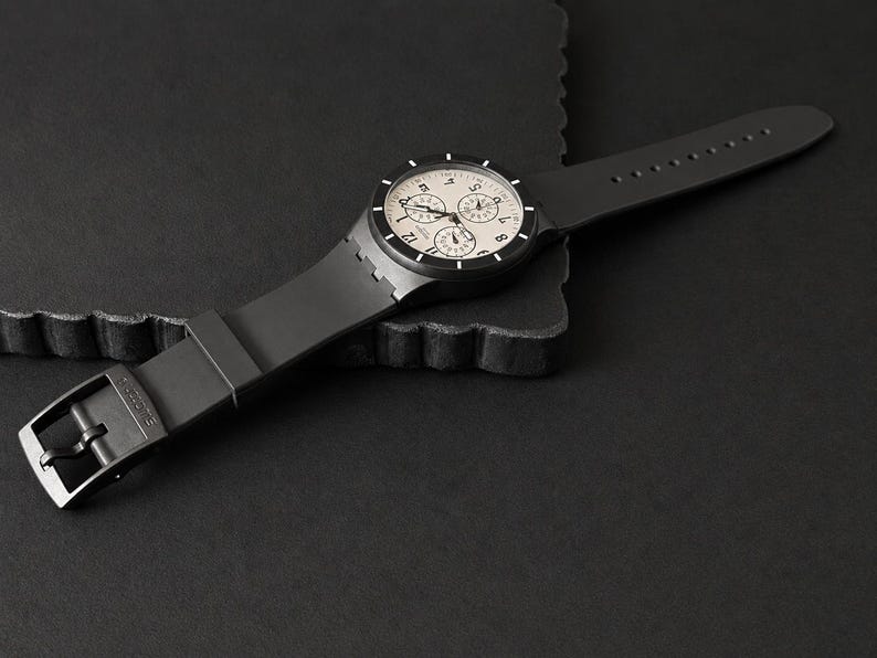 K&ouml;nnte beinhalten: Eine schwarze Armbanduhr mit hellem Zifferblatt und drei Hilfszifferbl&auml;ttern. Die Uhr hat ein schwarzes Armband und einen Schnallenverschluss. Die Uhr wird auf einer dunklen Oberfl&auml;che pr&auml;sentiert.