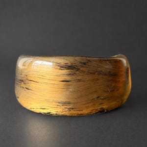 May include: A wide, semi-translucent cuff bracelet featuring a warm amber-toned base with dark, horizontal, brush-like streaks throughout the surface. The bracelet has a smooth, polished finish and a curved, open-ended design.