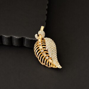 May include: A gold-toned leaf-shaped brooch with a textured surface. The brooch features a section of clear, sparkling crystals and a detailed, open-work design. The brooch is set against a black background.