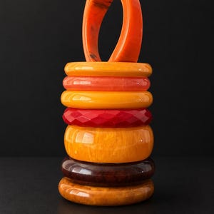 May include: A vertical stack of eight circular bangles in various shades of orange, yellow, red, and brown. The bracelets feature smooth, marbled, and faceted textures, with one orange bangle positioned upright at the top of the colorful collection.