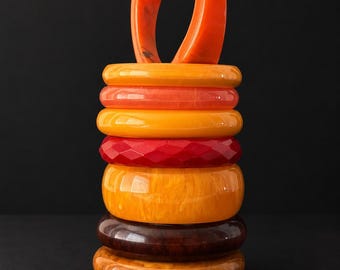 May include: A vertical stack of eight circular bangles in various shades of orange, yellow, red, and brown. The bracelets feature smooth, marbled, and faceted textures, with one orange bangle positioned upright at the top of the colorful collection.