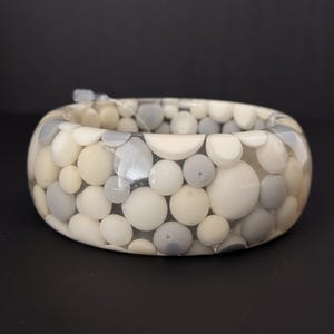 May include: A wide bangle bracelet featuring a clear resin base embedded with numerous smooth, circular beads in varying shades of cream, off-white, and light grey. The beads are arranged in a dense, multi-layered pattern throughout the band.