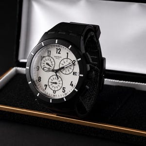 K&ouml;nnte beinhalten: Eine schwarze Swatch-Uhr mit wei&szlig;em Zifferblatt und schwarzer L&uuml;nette, pr&auml;sentiert in einer Pr&auml;sentationsbox. Die Uhr verf&uuml;gt &uuml;ber mehrere Hilfszifferbl&auml;tter und ein schwarzes Armband. Das Zifferblatt zeigt das Swatch-Logo.