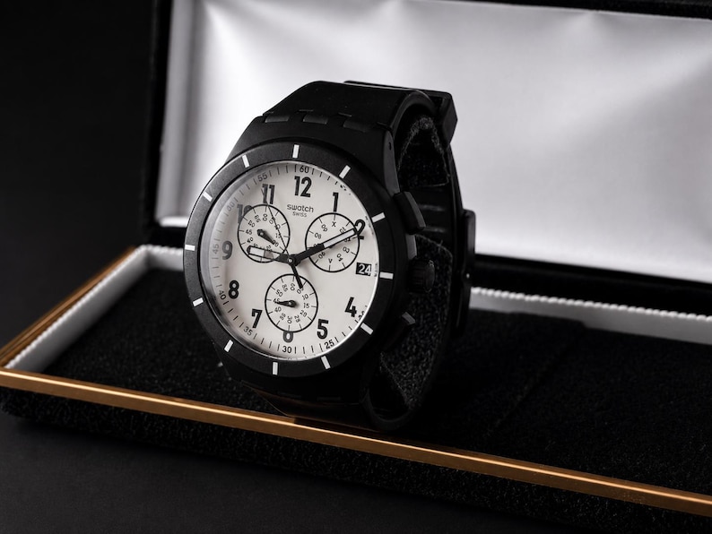 K&ouml;nnte beinhalten: Eine schwarze Swatch-Uhr mit wei&szlig;em Zifferblatt und schwarzer L&uuml;nette, pr&auml;sentiert in einer Pr&auml;sentationsbox. Die Uhr verf&uuml;gt &uuml;ber mehrere Hilfszifferbl&auml;tter und ein schwarzes Armband. Das Zifferblatt zeigt das Swatch-Logo.