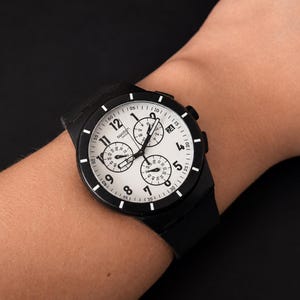 K&ouml;nnte beinhalten: Schwarze Armbanduhr mit wei&szlig;em Zifferblatt und schwarzer L&uuml;nette. Die Uhr verf&uuml;gt &uuml;ber drei Hilfszifferbl&auml;tter und ein schwarzes Armband. Die Uhr befindet sich an einem Handgelenk.