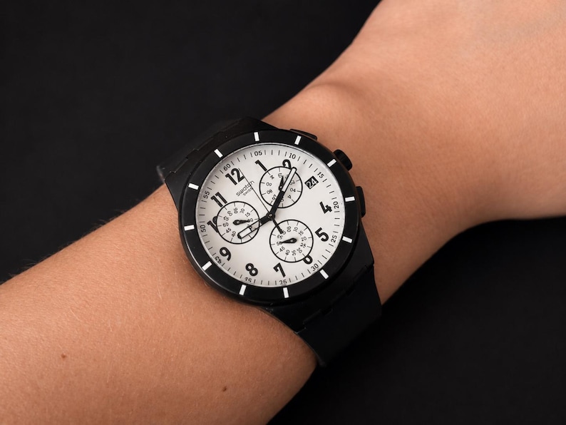 K&ouml;nnte beinhalten: Schwarze Armbanduhr mit wei&szlig;em Zifferblatt und schwarzer L&uuml;nette. Die Uhr verf&uuml;gt &uuml;ber drei Hilfszifferbl&auml;tter und ein schwarzes Armband. Die Uhr befindet sich an einem Handgelenk.