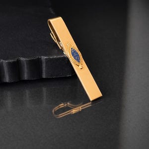 May include: A gold-colored tie bar with a blue and gold emblem. The tie clip is rectangular with a hinged clasp. The emblem has the word "THURST" in blue lettering. The tie bar is displayed on a dark surface.
