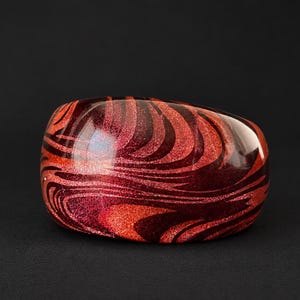 May include: A wide, smooth bangle bracelet featuring a swirling, marbled pattern of deep burgundy, bright red, and orange tones. The surface has a high-gloss, reflective finish with embedded glitter particles that catch the light across the curved form.