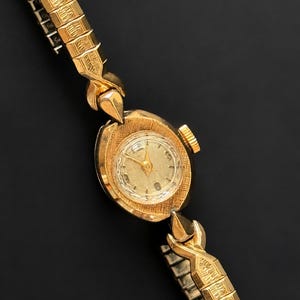 May include: A vintage gold wristwatch with a textured oval face and a gold band. The watch face has a clear cover and gold hands. The band is made of small, rectangular gold links. The watch is set against a black background.