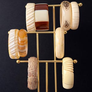 May include: A selection of ivory, brown, and amber-coloured bangles on a gold-toned metal stand. The bangles display diverse designs, including carved patterns, inlaid sections, and feather accents. The stand is set against a dark background.