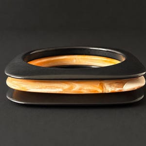 May include: A stack of three bangles featuring two solid matte black square-shaped bracelets and one central marbled amber and cream-coloured bracelet. The bangles are arranged horizontally, showcasing their smooth, rounded edges and contrasting colours.
