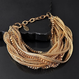 May include: Gold-coloured multi-strand bracelet. The bracelet features various chain styles, including box and curb chains, connected to a clasp. The jewellery is displayed on a black surface.