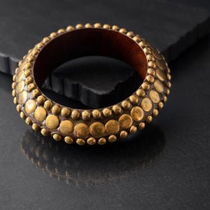 Peut inclure: Un bracelet doré avec un intérieur marron foncé. Le bracelet est orné de nombreux petits ornements circulaires dorés. Le fond est une surface sombre et texturée, rehaussant l'attrait visuel du bracelet. Un accessoire de déclaration.