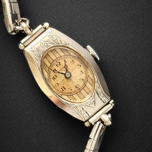 May include: An antique gold-toned wristwatch with an oval face and a silver-toned band. The watch face features a light-colored background with black numerals and hands. The watch case has decorative engravings. The band is made of small, rectangular links.