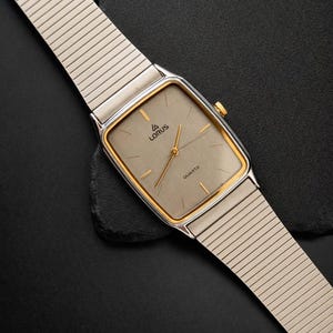 May include: A silver-toned Lorus quartz watch with a rectangular face and gold-colored trim. The watch has a light gray dial with gold hands and the word "QUARTZ" printed on the face. The watch band is a silver-toned, flexible metal mesh.