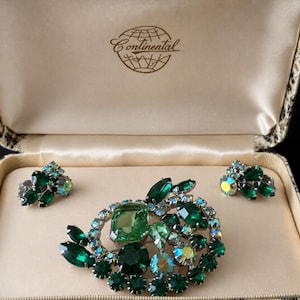 Puede incluir: Un conjunto de joyería que incluye un broche y dos pendientes a juego, adornados con pedrería verde e iridiscente sobre metal plateado. El set se presenta en una caja de color crema con el texto Continental impreso en el interior.