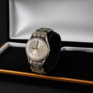 May include: A clear wristwatch with a silver dial and green accents on the band, displayed in a black box with a gold trim. The watch has a transparent plastic band and a silver face with black hands.