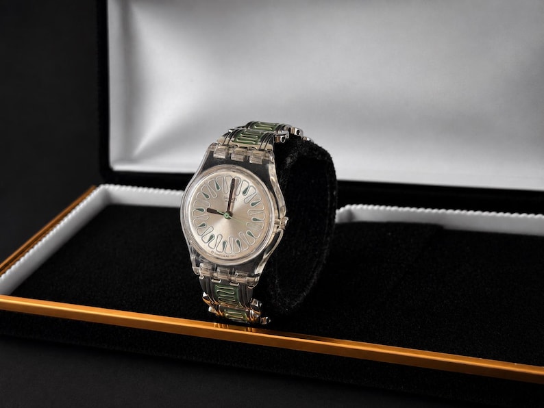 May include: A clear wristwatch with a silver dial and green accents on the band, displayed in a black box with a gold trim. The watch has a transparent plastic band and a silver face with black hands.