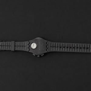 K&ouml;nnte beinhalten: Eine schwarze Armbanduhr mit schwarzem Armband, Schnalle und Zifferblatt. Die Uhr liegt flach auf einem schwarzen Hintergrund. Das Zifferblatt hat einen silberfarbenen Knopf.