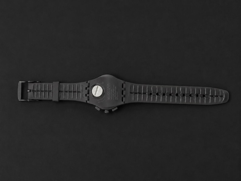 K&ouml;nnte beinhalten: Eine schwarze Armbanduhr mit schwarzem Armband, Schnalle und Zifferblatt. Die Uhr liegt flach auf einem schwarzen Hintergrund. Das Zifferblatt hat einen silberfarbenen Knopf.