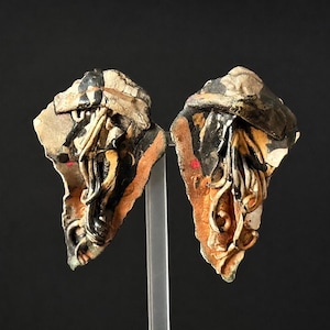 May include: Two abstract ceramic sculptures, each with a unique shape and texture. The pieces feature a mix of colors, including black, tan, and orange, with intricate details. The sculptures are displayed against a dark background.