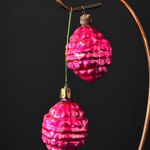 May include: Two vintage glass Christmas ornaments in a vibrant magenta pink color with a textured, grid-like pattern resembling pinecones. Each ornament features a metallic silver-toned cap and hangs from a thin green string on a curved metal stand.