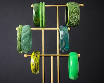 May include: A gold-colored jewelry display stand holding various green bangles. The bangles vary in shades and designs, including solid colors, floral carvings, and patterned textures. The background is a dark, solid color.