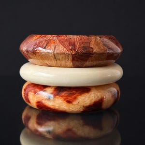 May include: Three stacked wooden bangles. The top bangle is a mosaic of reddish-brown wood pieces. The middle bangle is a solid cream color. The bottom bangle has a mottled pattern of reddish-brown and tan.